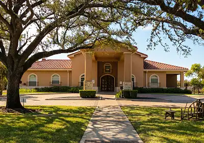 Beige stucco building with a red tile roof and arched entrance, approached by a central brick walkway flanked by trees and a bench on the right.