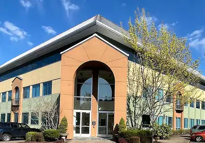 Modern two-story office building with a large arched entryway and glass doors, surrounded by shrubs and trees under a blue sky.”,
