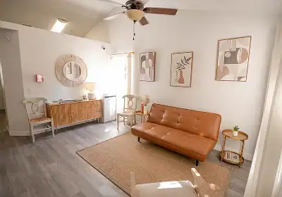 Bright living room featuring a tan leather sofa, light wood coffee table, beige rug, wall art, ceiling fan, round mirror, and a sideboard with chairs.