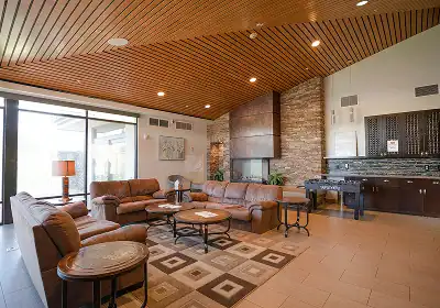 Contemporary hotel lobby: brown leather sofas around circular coffee tables, with a brick fireplace and wood-paneled ceiling; kitchenette along the back wall.