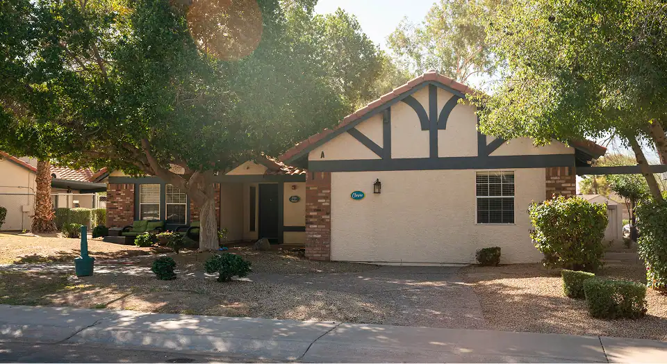 Front view of a single-story ranch-style house with beige stucco, dark trim, brick accents, and a large tree in the yard under sunny skies.