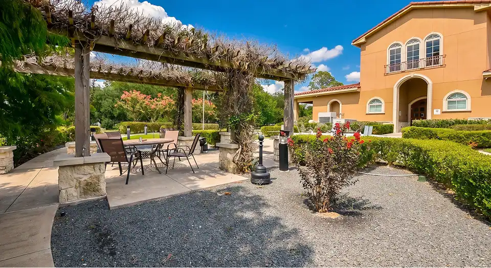 Outdoor patio area with a wooden pergola casting shade over a metal table and chairs, nearby stone pillars and manicured bushes by a tan stucco building with arched windows under a bright blue sky.