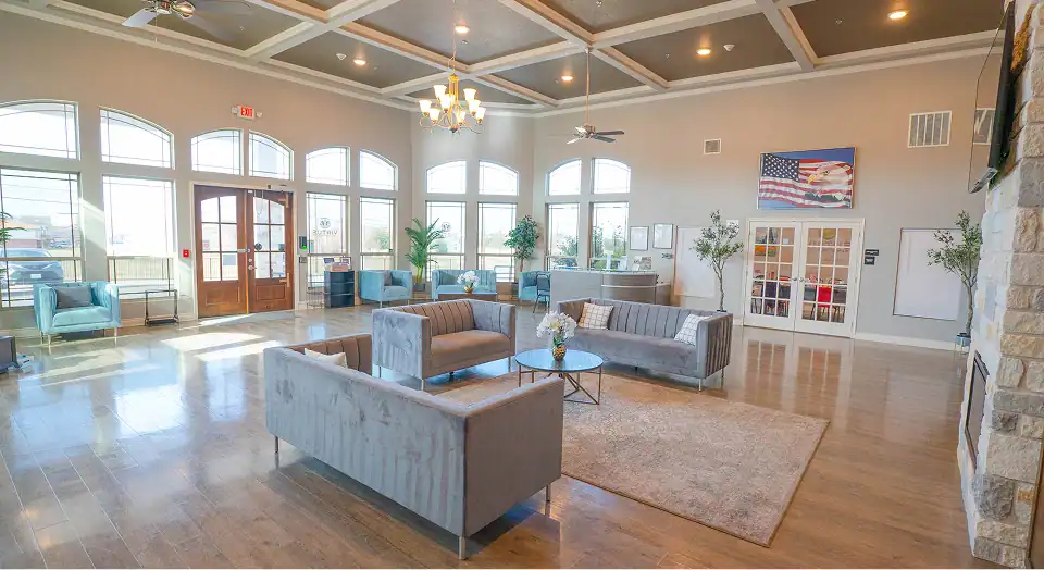 Spacious lobby with gray sofas arranged in a seating area, coffee tables, and a beige rug under wood floors; large arched windows and chandelier overhead.