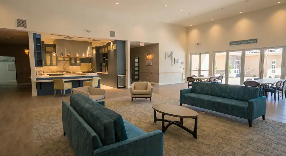 Spacious lobby with teal sofas around a coffee table, open kitchen bar to the left, and glass doors to an outdoor dining area on the right.