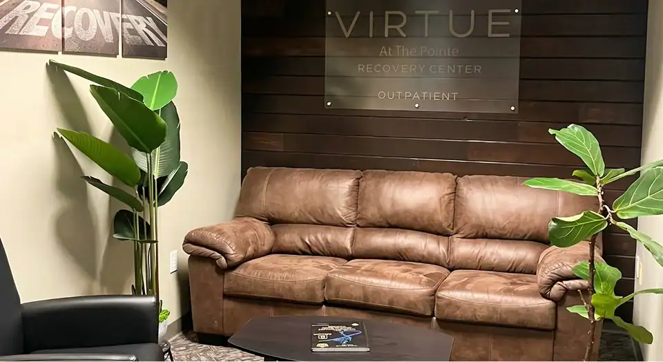 Brown leather sofa in a waiting room with a glass sign reading 'VIRTUE At The Pointe Recovery Center Outpatient' on a dark wood wall.