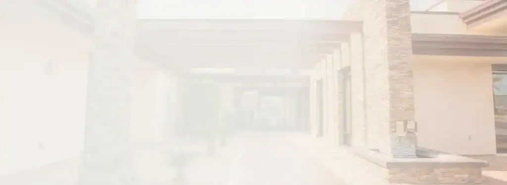 A sunlit walkway with stone walls, large windows, and a wooden pergola overhead, leading to an entrance; small plants line the path. The image is overexposed, creating a soft, washed-out effect.