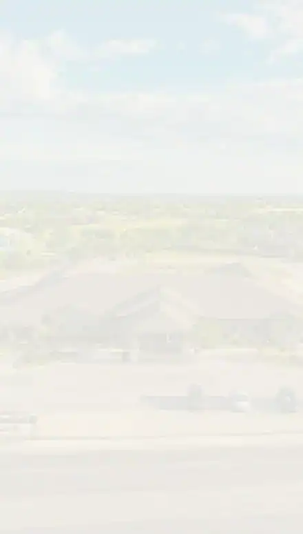 A lightly faded aerial view of a suburban area with a large building, parking lot, green trees, and scattered houses under a partly cloudy sky.