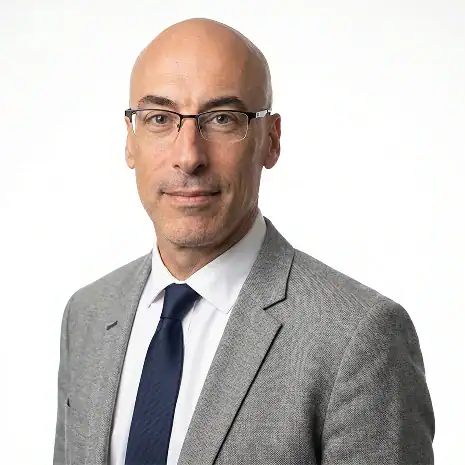 A man with glasses, a bald head, and light skin is wearing a light gray suit, white shirt, and dark blue tie. He is facing forward and smiling slightly against a plain white background.