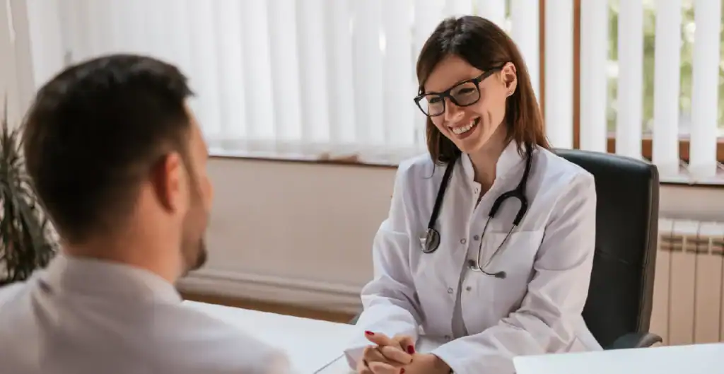 A female doctor wearing glasses and a lab coat smiles while sitting at her desk, speaking with a male patient in a bright, modern office.