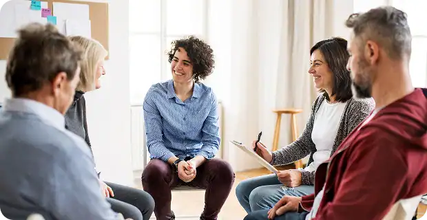 Five adults sit in a circle indoors, engaged in conversation. One person holds a clipboard and pen, possibly leading the discussion. The group appears relaxed and attentive in a bright room with large windows.