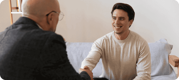Two men shake hands and smile, seated on a sofa, indicating a friendly meeting or agreement.