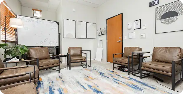 Waiting room with six brown leather chairs, glass coffee tables, a whiteboard, and plants in a bright office space.