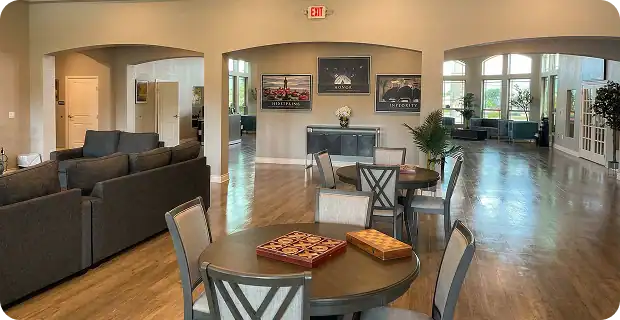A spacious, well-lit lounge with round tables and chairs, board games on the tables, gray couches, wooden floors, large windows, and framed artwork on the walls.