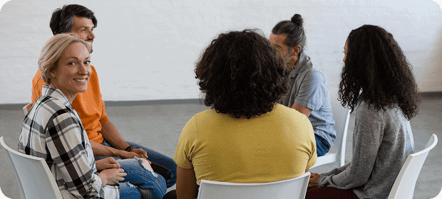 Six adults sit in a circle on white chairs in a bright room, engaged in a discussion; a woman in a plaid shirt smiles toward the camera.