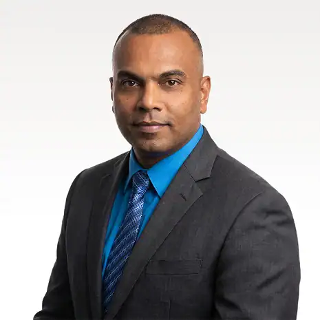 A man in a gray suit, blue dress shirt, and blue patterned tie poses against a plain light background, looking confidently at the camera.