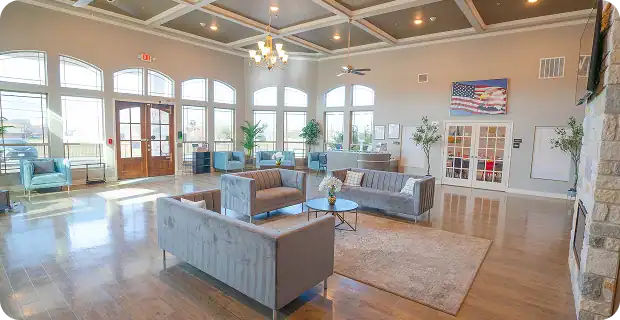 Spacious, bright lobby with large windows, gray sofas, glass coffee tables, potted plants, and an American flag on the wall. Light wood flooring and a chandelier complete the modern, welcoming space.