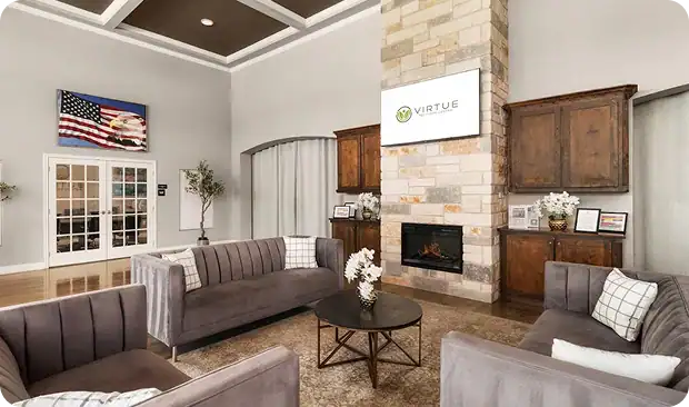 A modern lobby with gray sofas, a round coffee table, a stone fireplace, and a TV displaying the Virtue logo. An American flag hangs on the wall above glass doors; wooden cabinets and flower arrangements add decor.