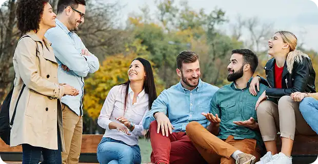 Six young adults are sitting and standing outdoors on a bench, smiling and laughing together. The background shows trees with autumn leaves, creating a warm and relaxed atmosphere.
