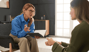 A therapist with glasses and a blue shirt sits with an open notebook, attentively listening and talking to a person seated across from her in an office setting.