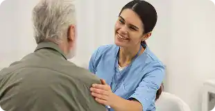 Smiling nurse in blue scrubs comforting an elderly man with a hand on his shoulder in a clinical setting.