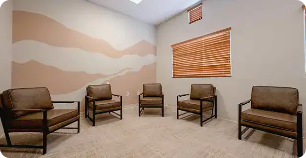 Waiting room with five brown cushioned chairs arranged in a loose circle, beige walls with a peach wavy mural, and a window with wooden blinds nearby.—Informative and contextual for a waiting area image.