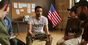 Man wearing a white ARMY shirt speaks to a small group seated in a circle in a room with an American flag and bulletin board in the background.