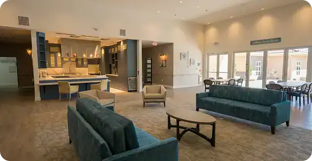 Bright, open lounge with two blue sofas around a wooden coffee table, beige chairs, and a kitchen area in the back; large windows and glass doors to an outdoor space.