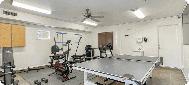 Home gym in a basement with a ping-pong table in the foreground, plus cardio machines, weights, and a ceiling fan in the background.