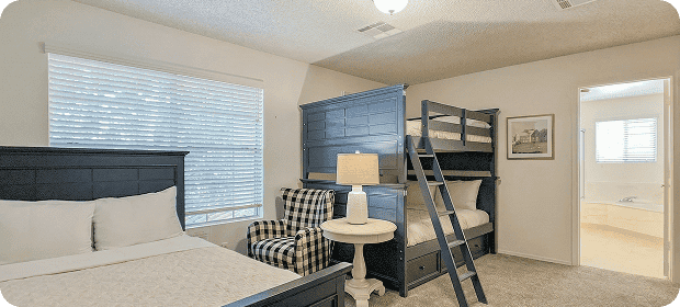 Spacious hotel room with a queen bed in front and a blue bunk bed against the wall, a checkered chair, round white side table with a lamp, large window with blinds, carpeted floor, and a doorway to a bathroom.