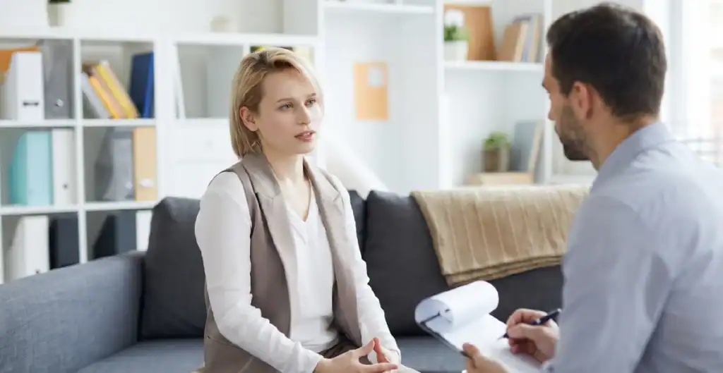 A woman sits on a couch talking to a man holding a clipboard, possibly in a therapy or counseling session, in a bright, modern office with shelves and decor in the background.