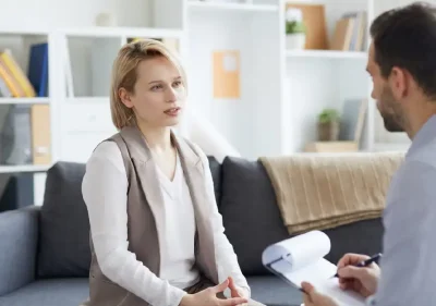 A woman sits on a couch talking to a man holding a clipboard, possibly in a therapy or counseling session, in a bright, modern office with shelves and decor in the background.