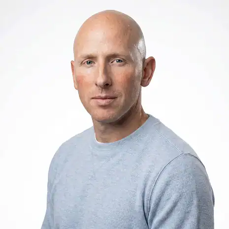 A bald man wearing a light gray long-sleeve shirt looks at the camera with a neutral expression against a plain white background.