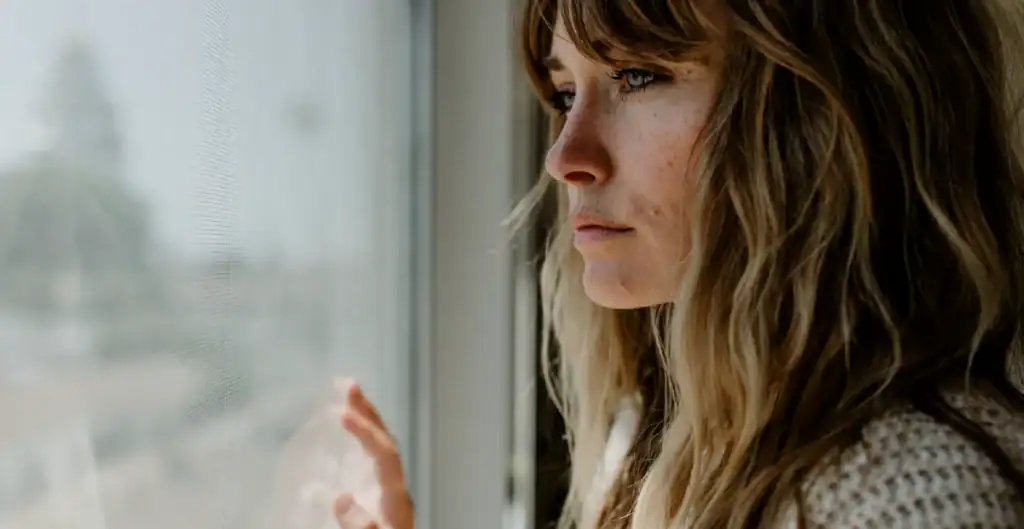 A woman with long, wavy hair looks pensively out a window, touching the glass with her hand. Soft daylight illuminates her thoughtful expression and the side of her face.