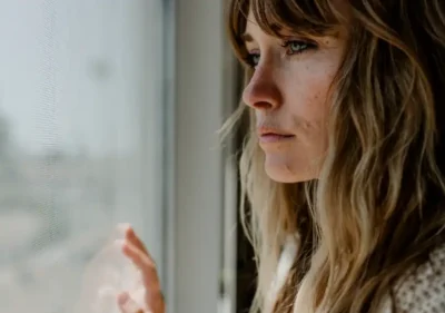 A woman with long, wavy hair looks pensively out a window, touching the glass with her hand. Soft daylight illuminates her thoughtful expression and the side of her face.