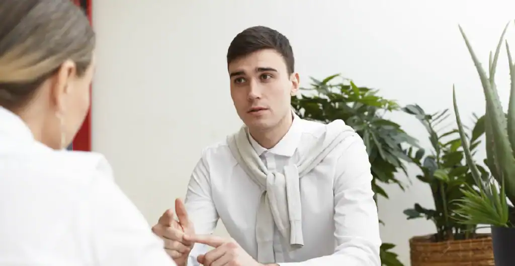 A young man in a white shirt and sweater talks seriously with a woman across a table, surrounded by green plants in a bright room.