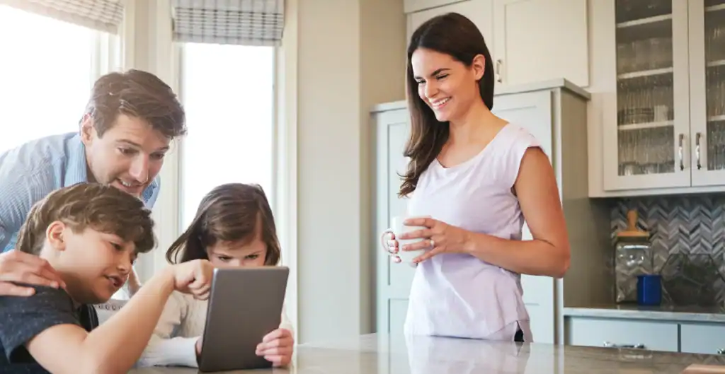 A woman stands smiling with a mug in hand while a man and two children look at a tablet together at a kitchen counter, with sunlight streaming through the windows.