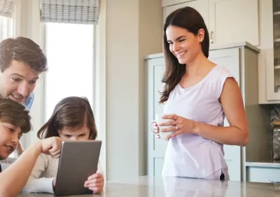 A woman stands smiling with a mug in hand while a man and two children look at a tablet together at a kitchen counter, with sunlight streaming through the windows.