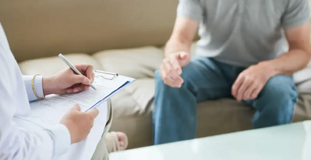 A person sitting on a couch talks to a professional holding a clipboard and pen, taking notes during a counseling or therapy session. Only their torsos and hands are visible.