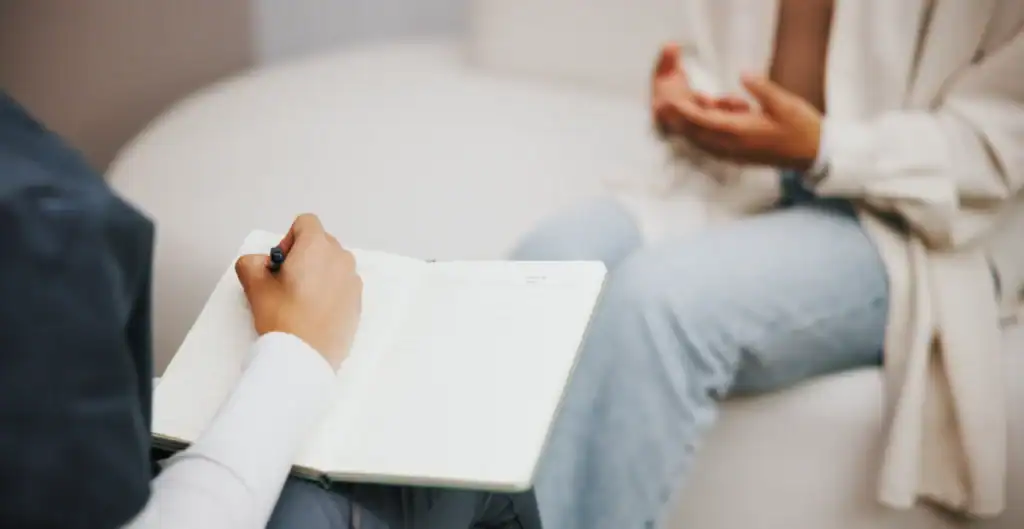 A person holding a notebook and pen takes notes while another person sits across from them, gesturing with their hands during a conversation. Both individuals are partially visible, seated in a casual setting.