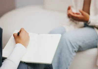 A person holding a notebook and pen takes notes while another person sits across from them, gesturing with their hands during a conversation. Both individuals are partially visible, seated in a casual setting.