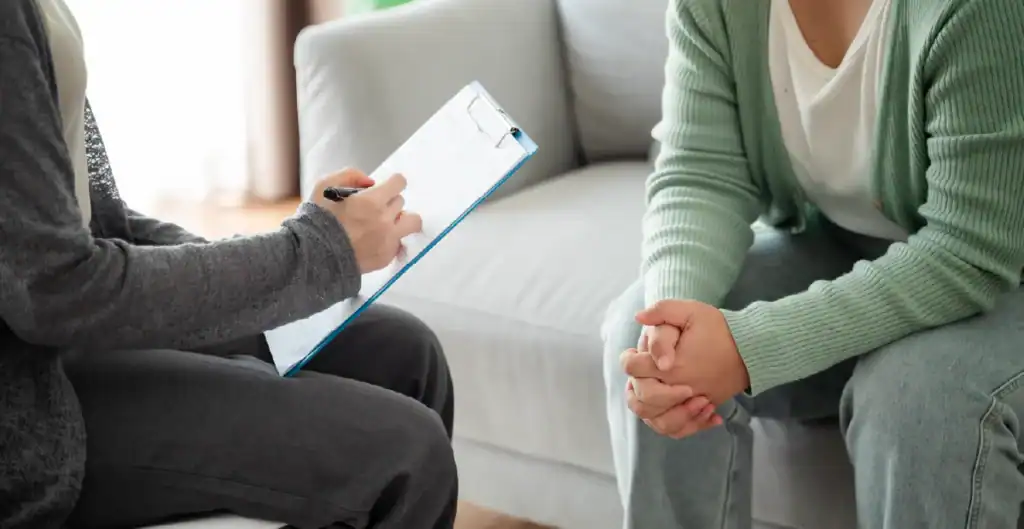Two people sit on a couch during a counseling session; one holds a clipboard and pen, while the other sits with hands clasped, suggesting a conversation or interview in a comfortable setting.