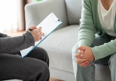 Two people sit on a couch during a counseling session; one holds a clipboard and pen, while the other sits with hands clasped, suggesting a conversation or interview in a comfortable setting.