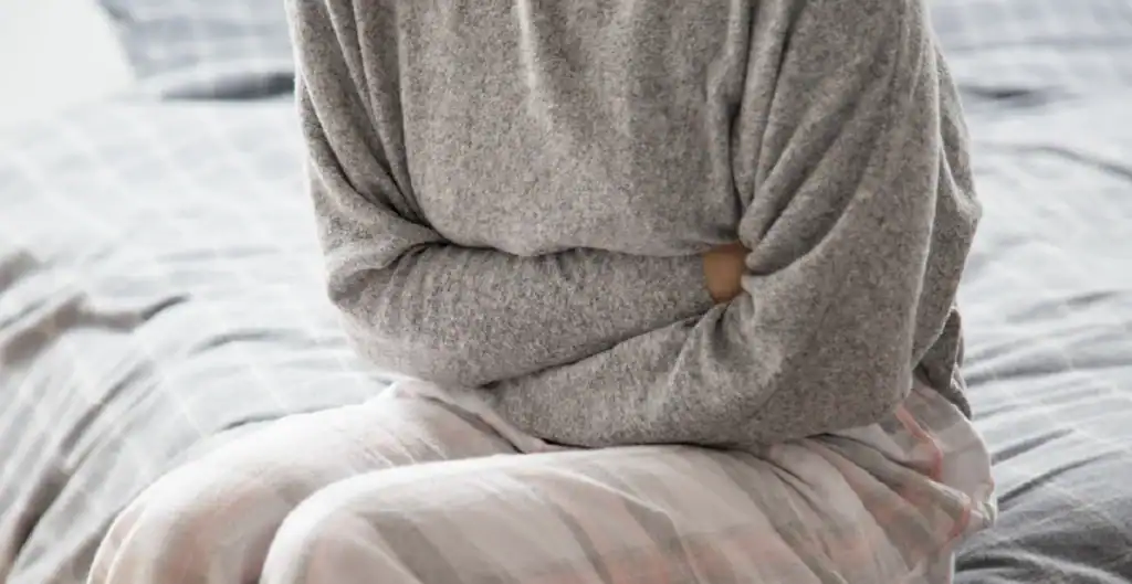 A person sitting on a bed, wearing a gray sweater and plaid pants, is holding their stomach as if experiencing discomfort or pain.