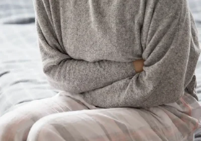 A person sitting on a bed, wearing a gray sweater and plaid pants, is holding their stomach as if experiencing discomfort or pain.