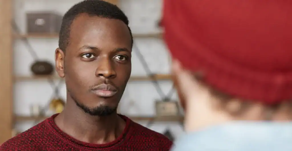 A man in a red sweater looks seriously at another person wearing a red beanie, who is facing away from the camera. The background is blurred, showing shelves with various objects.