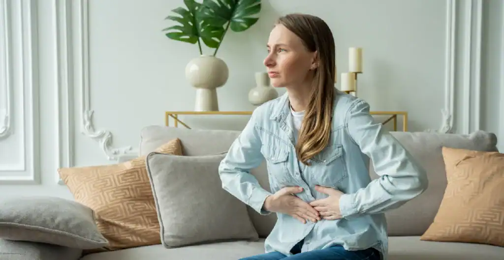 A woman sits on a couch with her hands pressed against her stomach, appearing to be in discomfort. She wears a light denim shirt and there are decorative pillows and plants in the background.