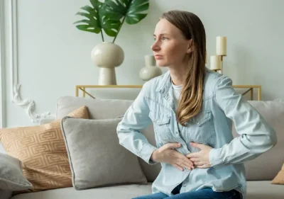 A woman sits on a couch with her hands pressed against her stomach, appearing to be in discomfort. She wears a light denim shirt and there are decorative pillows and plants in the background.