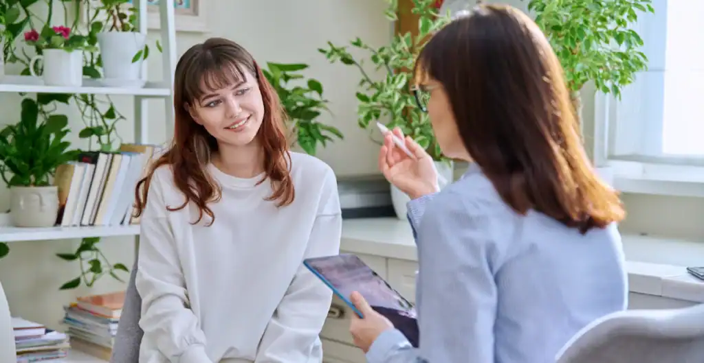 A young woman sits and smiles while talking to a professional holding a clipboard in a bright office filled with plants and books.