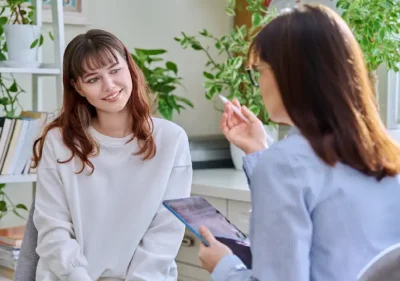 A young woman sits and smiles while talking to a professional holding a clipboard in a bright office filled with plants and books.