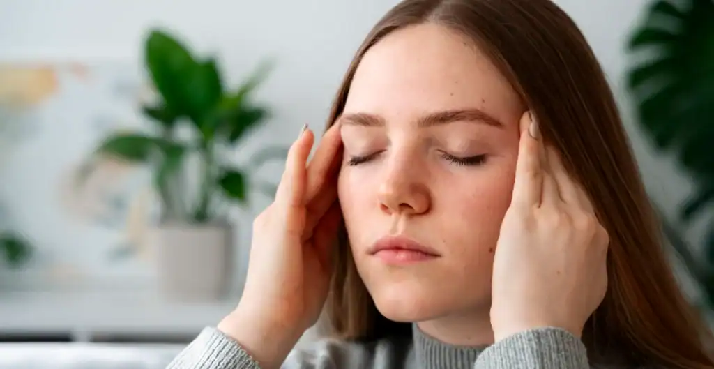 A young woman with closed eyes touches her temples with both hands, appearing to have a headache or feel stressed. There is a green leafy plant blurred in the background.